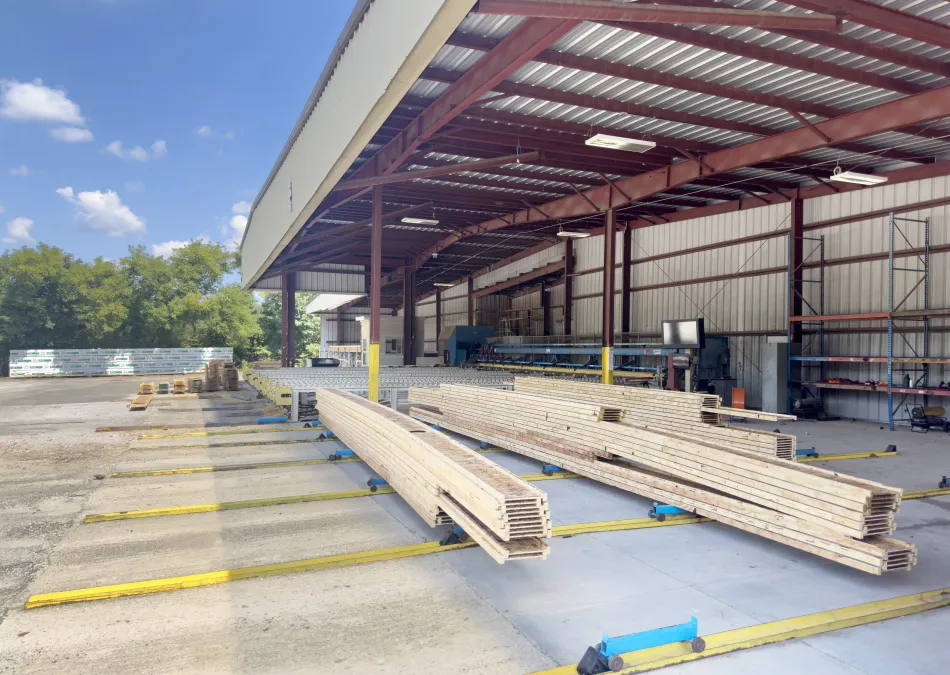 Wood planks stacked under a large metal-roofed warehouse with open sides on a sunny day.