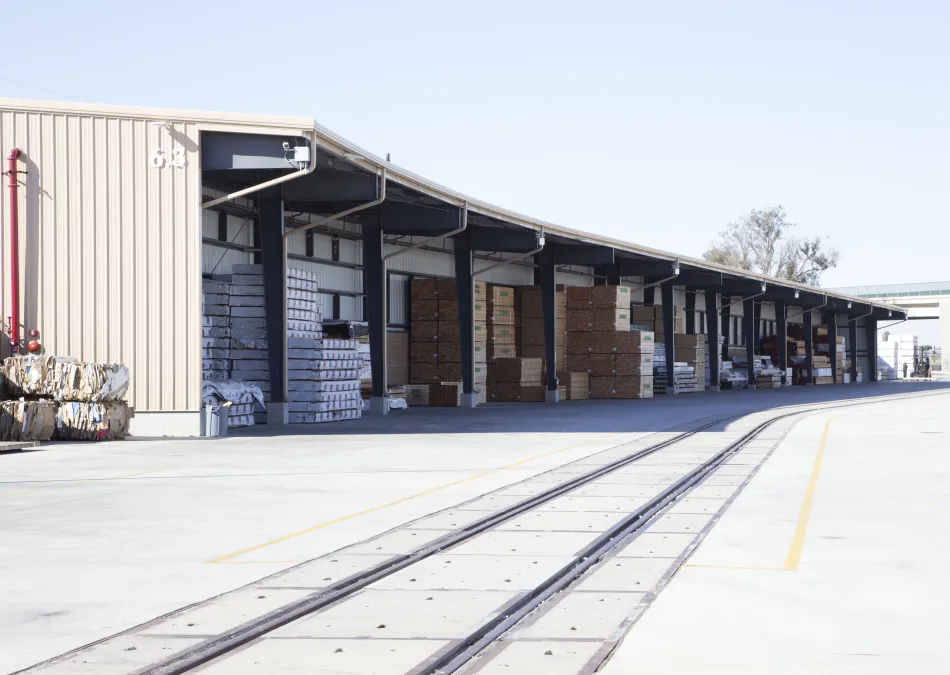 Outdoor warehouse storage with stacks of bundled packages and railway tracks in front under clear sky.