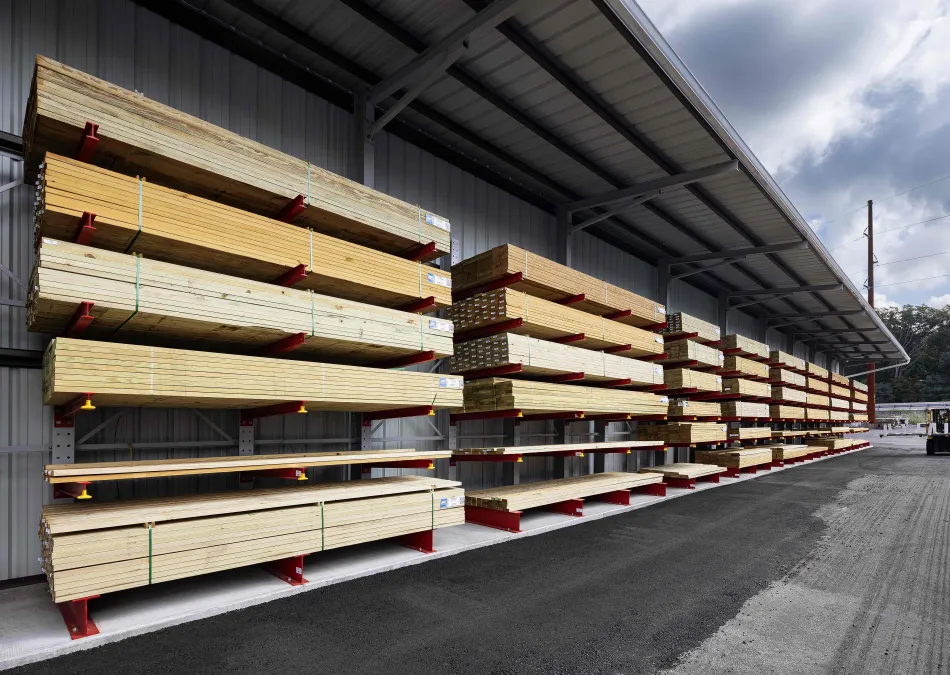 Long outdoor shelves stacked with bundles of lumber under a metal roof with a cloudy sky background.