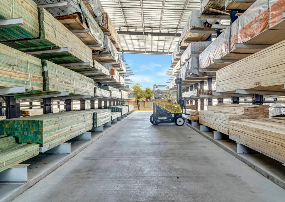 Lumber yard aisle with stacked wooden planks on metal racks and a yellow forklift under a metal roof.