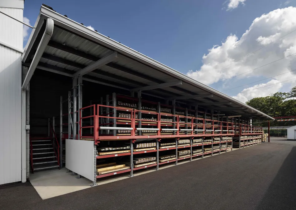 Outdoor warehouse racks with multiple shelves storing wooden planks under a metal roof on a sunny day.