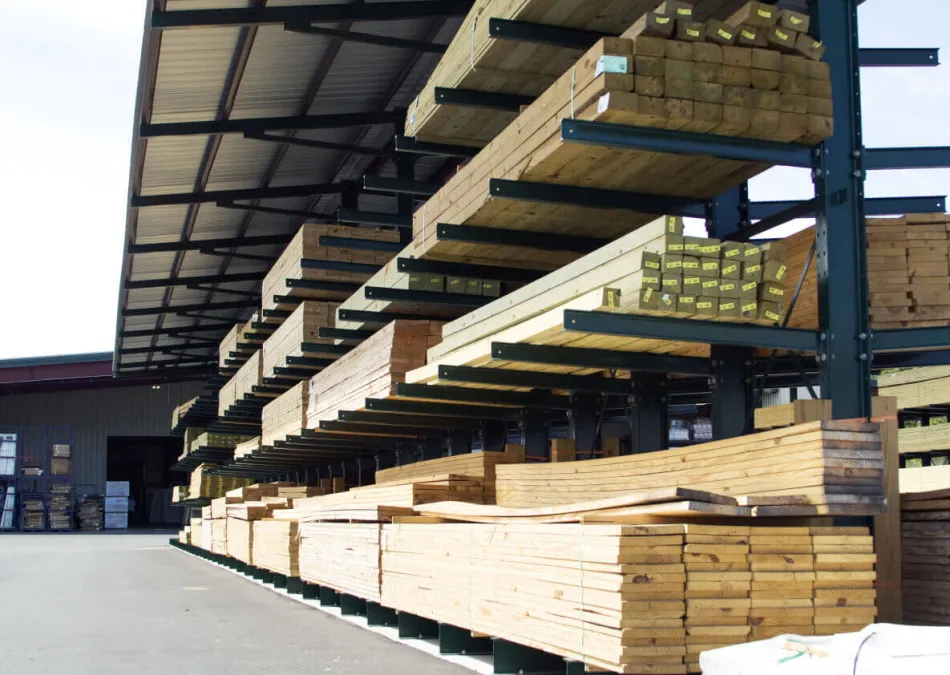 Outdoor lumber yard with neatly stacked wooden planks and beams under metal roofing on storage racks.
