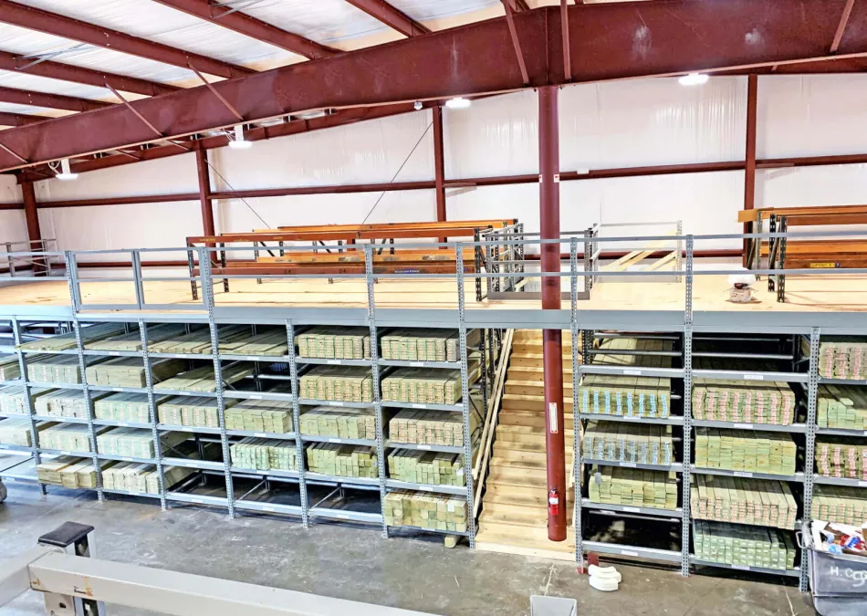 Warehouse with stacked wooden planks on metal shelves and a mezzanine level with wooden stairs and railings.