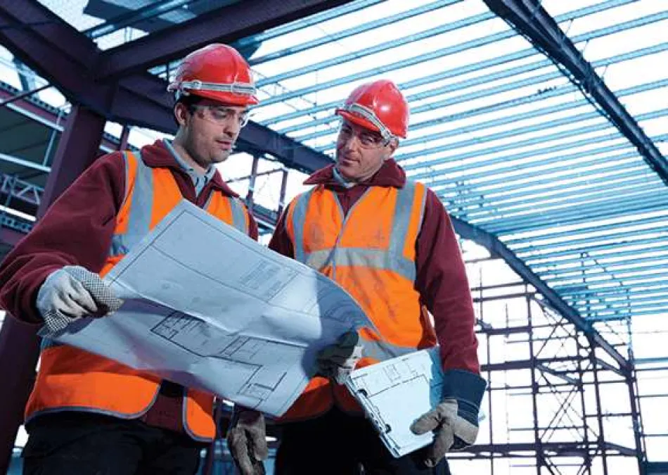Two construction workers in safety gear reviewing blueprints inside a steel-framed building under construction.