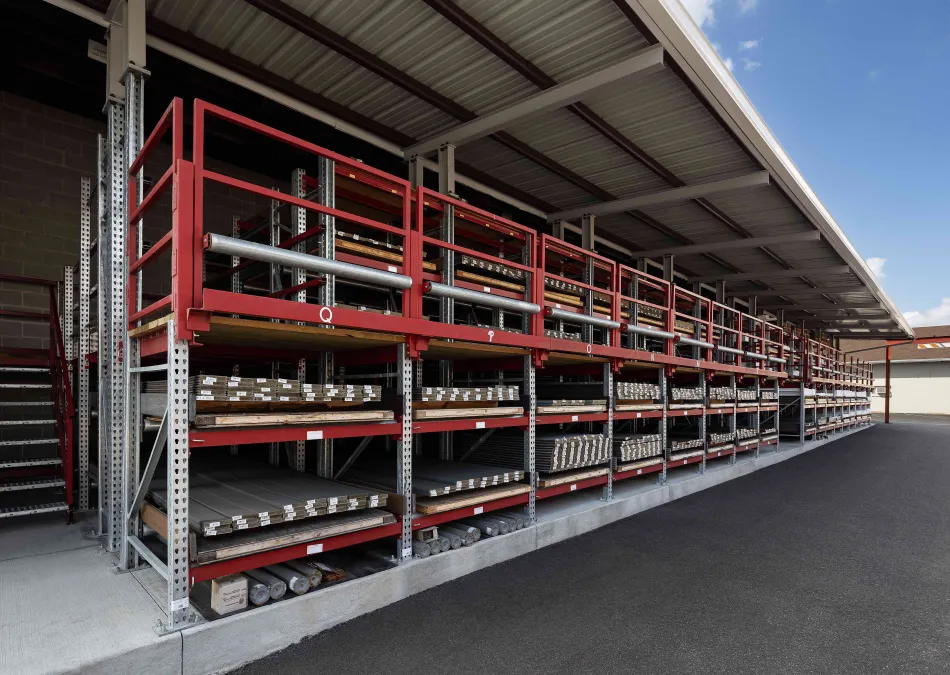 Outdoor industrial storage racks with red shelving holding metal pipes under a roof on a paved area.