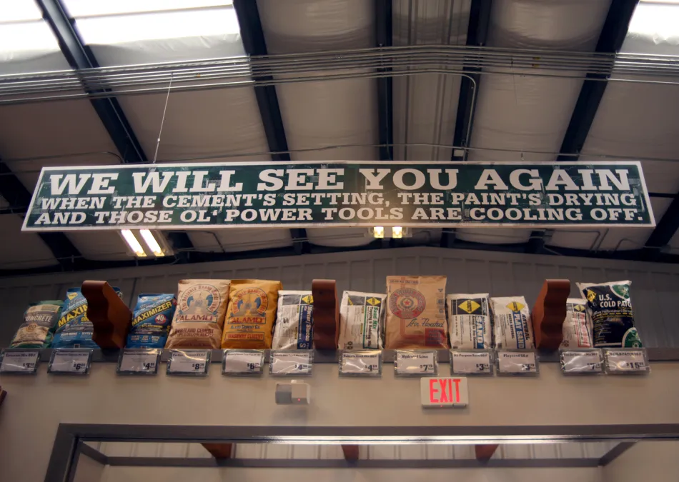 Sign in a hardware store reads We Will See You Again above various cement and powder bags with pricing.