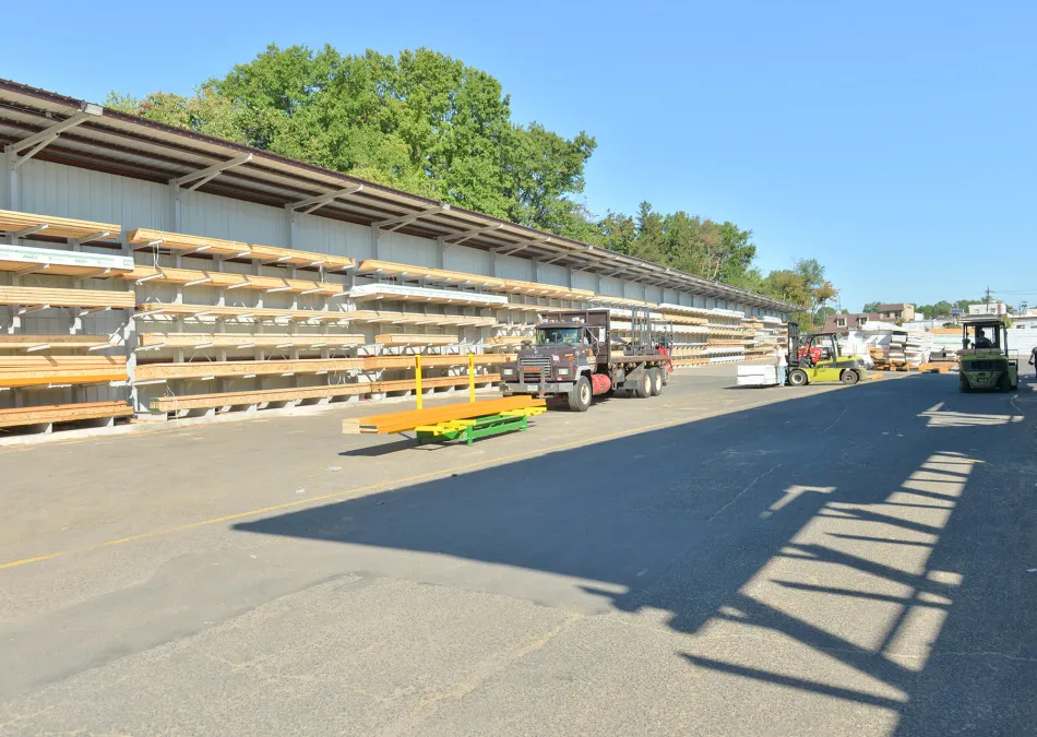 Outdoor lumber yard with stacked wood planks, trucks, forklifts, and clear blue sky on a sunny day.
