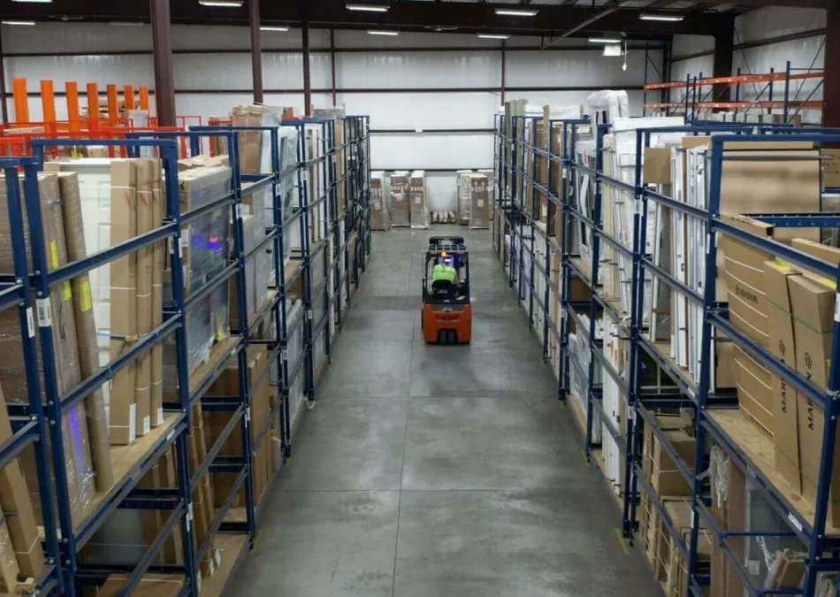 Warehouse aisles with metal racks filled with boxed goods and a forklift in the center aisle under bright lighting.