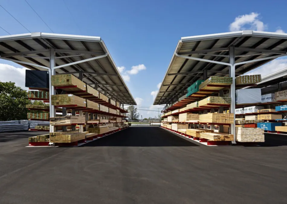 Outdoor lumber storage racks with various wood planks under metal roofs on a clear sunny day with blue sky.