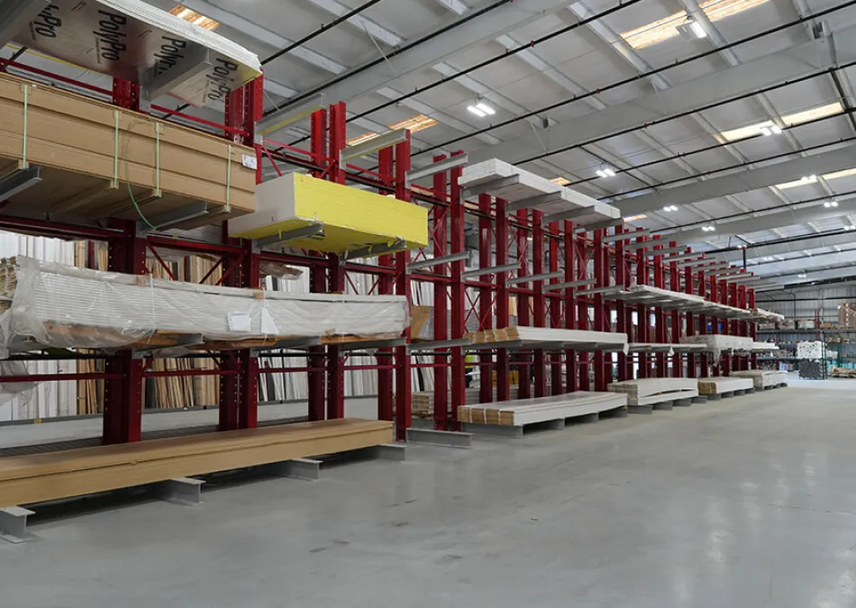 Spacious warehouse interior with red metal racks holding stacks of wood panels and construction materials.