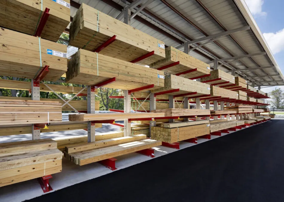 Shelves stocked with stacks of cut lumber under a metal roof in an outdoor storage area on a clear day