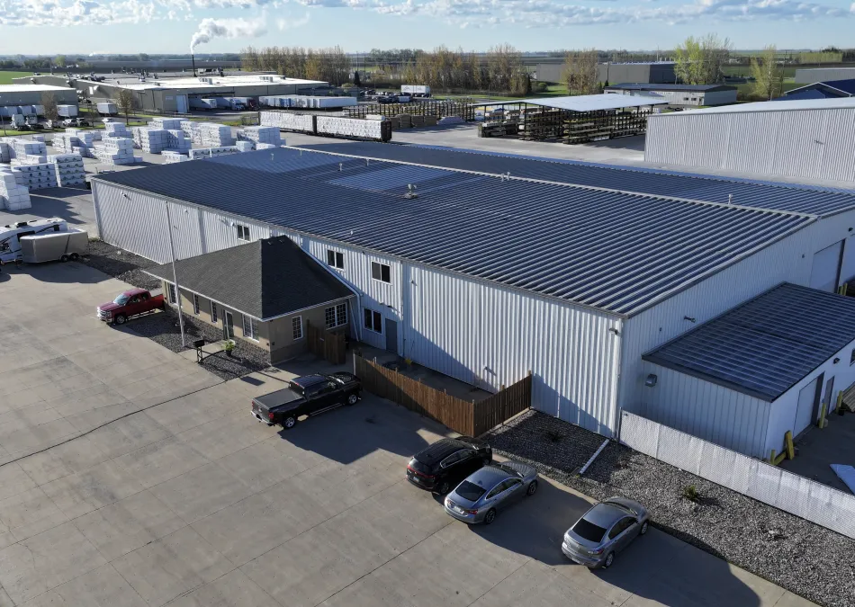 Aerial view of a large industrial warehouse with parked cars and stacks of packaged materials outside under a cloudy sky