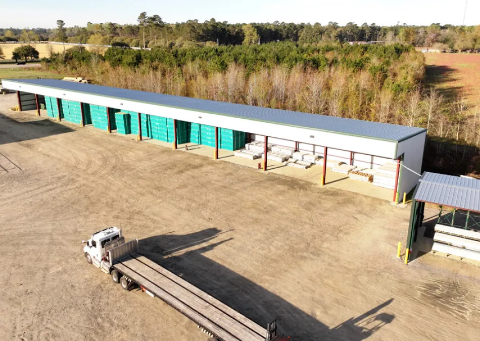Large industrial warehouse with green doors and a white semi-truck parked in a dirt lot on a sunny day