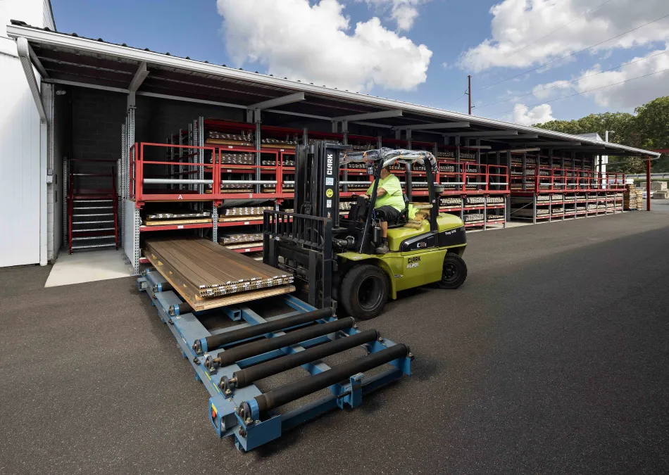 Forklift operator moving metal sheets on roller conveyor outside large warehouse with metal storage racks.