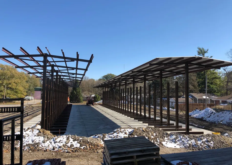 Construction site with steel frame structures and concrete foundation under clear blue sky, surrounded by trees and snow patches.