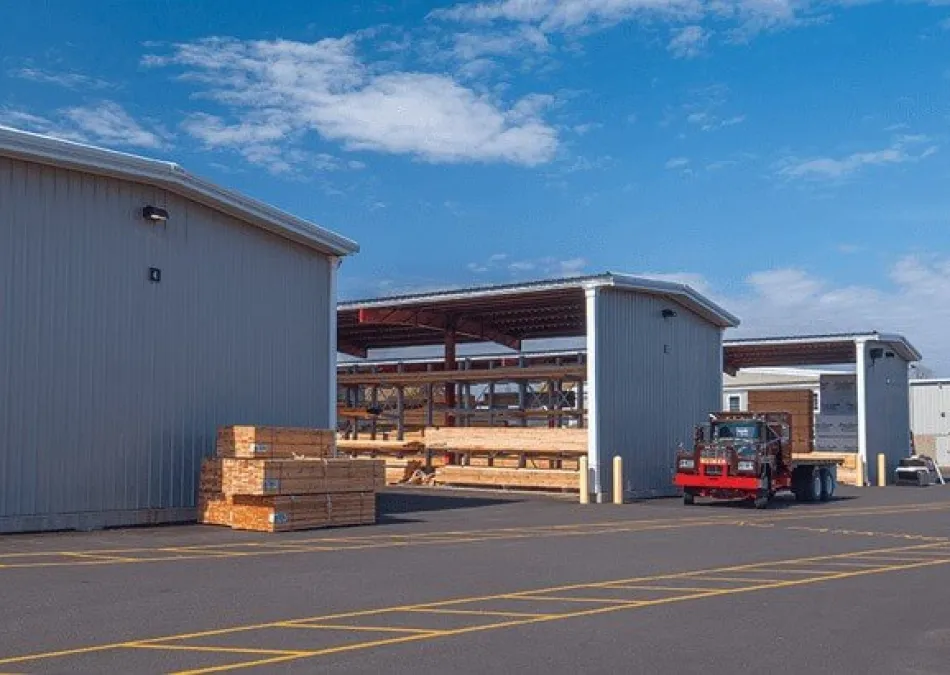Industrial lumberyard with stacked wood, metal storage buildings, and a red truck under a blue sky with clouds