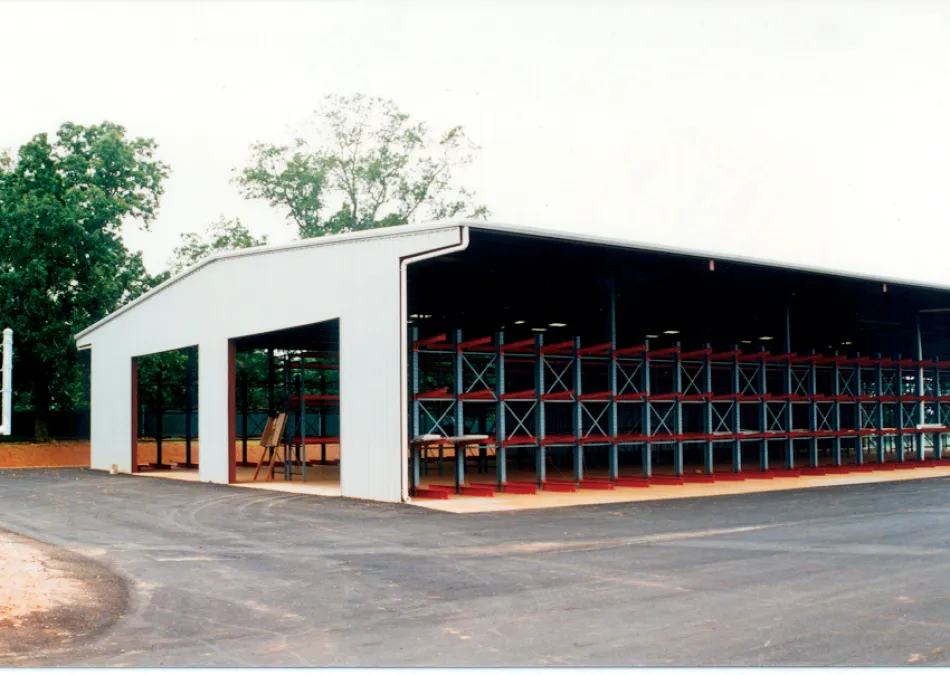 Large open warehouse with metal shelving units and paved driveway under cloudy sky near trees