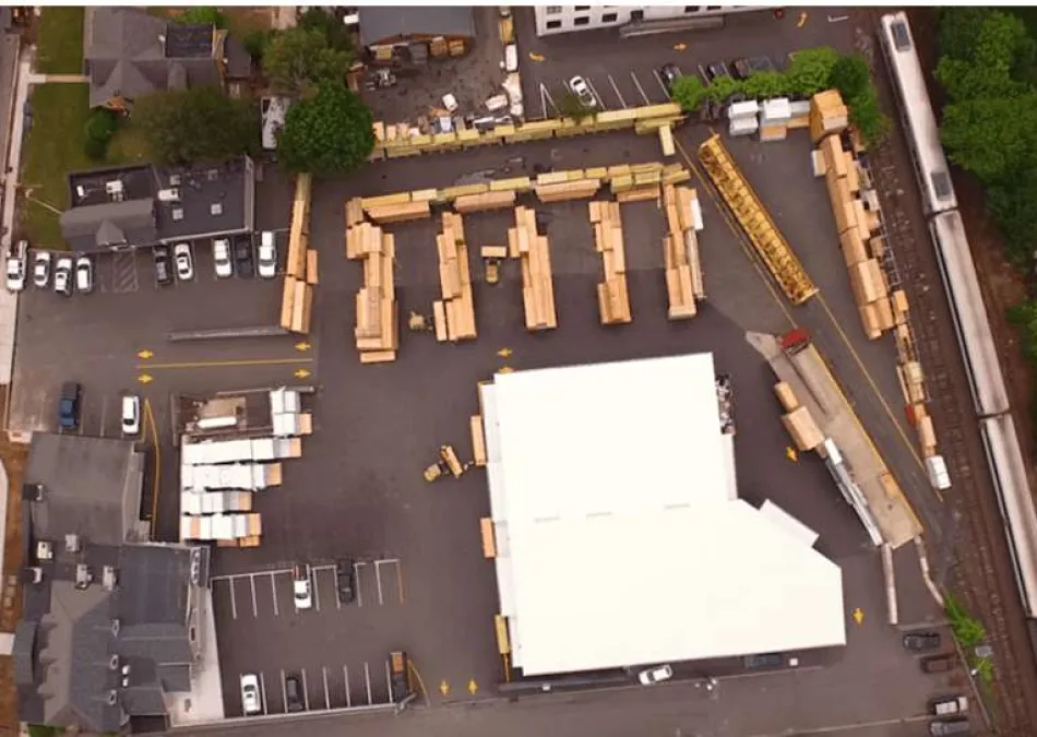 Aerial view of a warehouse with stacked wooden crates, surrounding buildings, parked cars, and nearby train tracks.