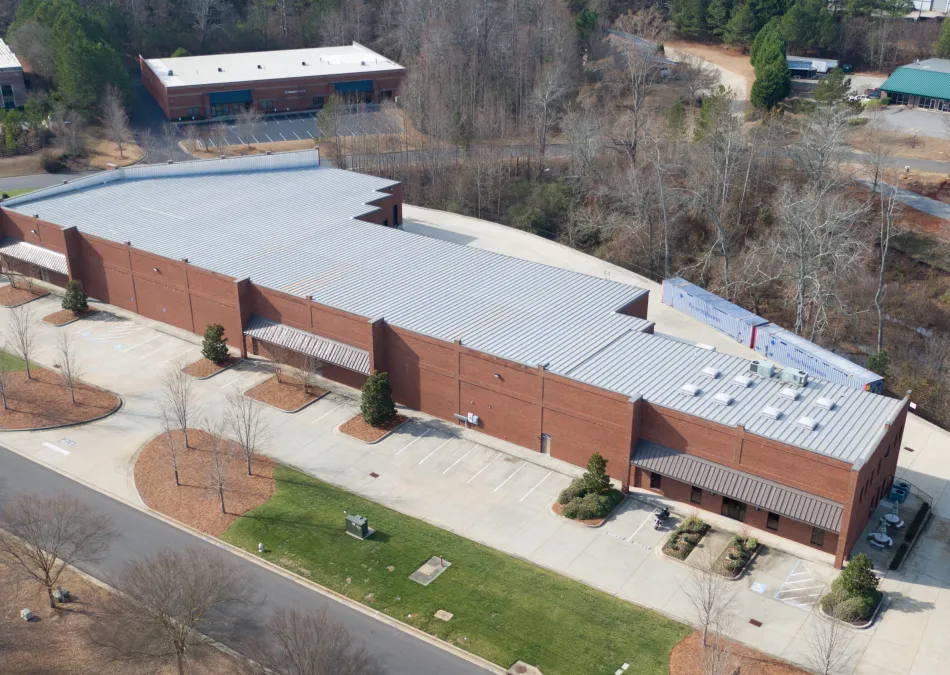Aerial view of a large brick commercial building with a gray metal roof surrounded by trees and parking spaces.