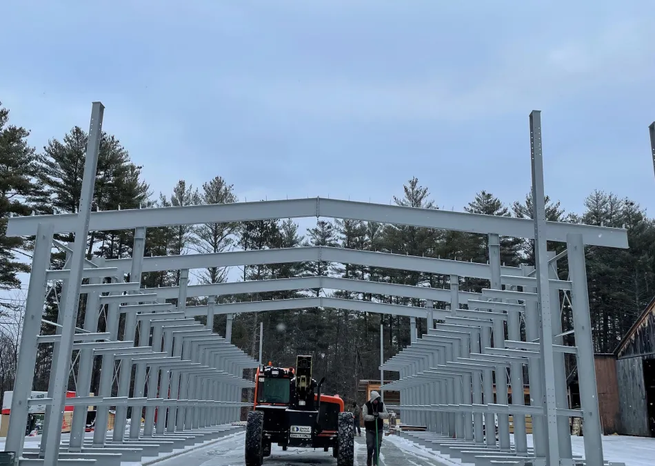 Steel structure framework under construction in snowy outdoor setting with trees and construction equipment.