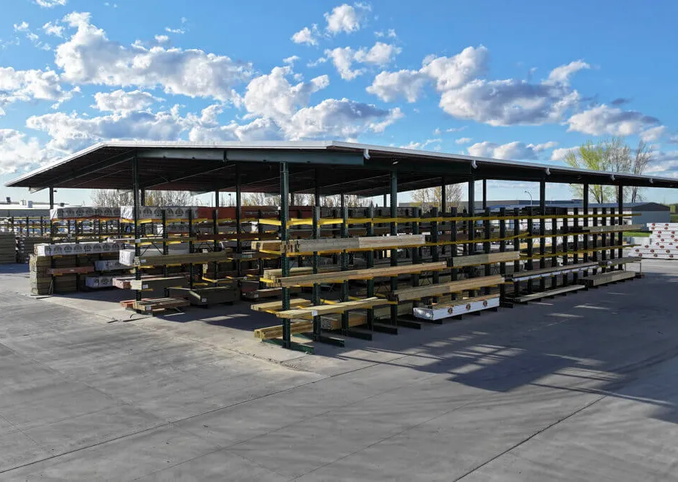 Outdoor lumber storage with metal racks under a large roof on concrete ground under a partly cloudy sky.