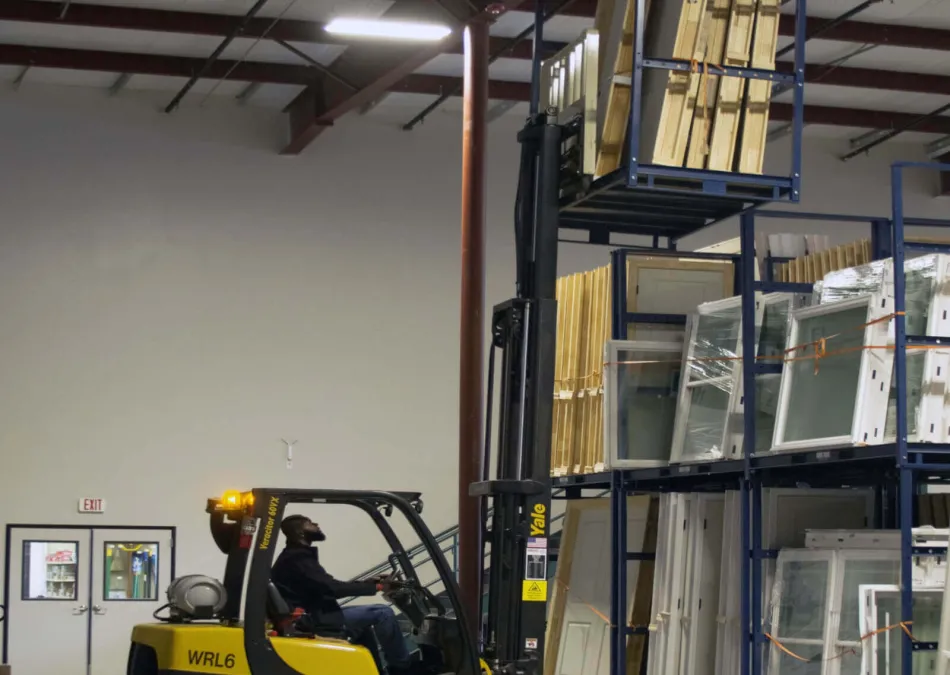 Worker operating yellow Yale forklift lifting wooden panels in a warehouse with metal shelving and various stored materials.
