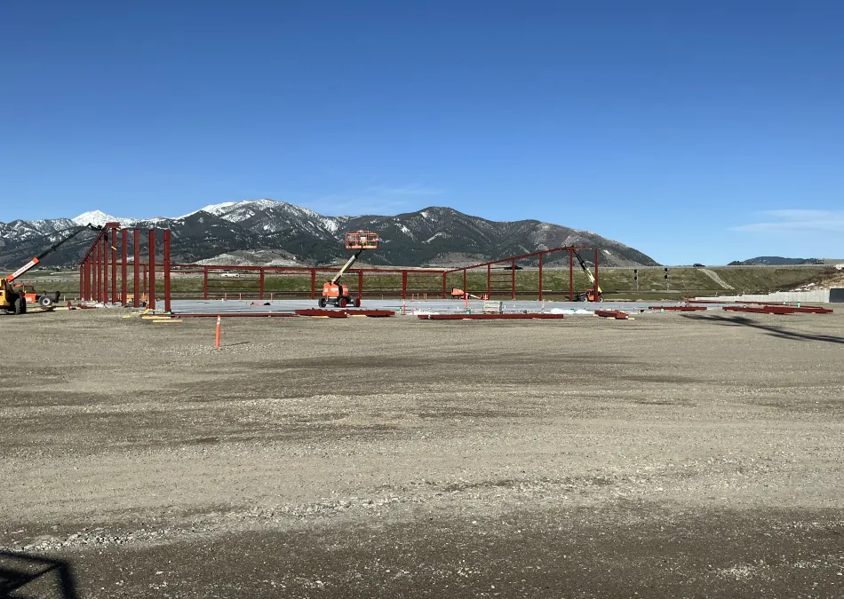 Construction site with red steel framework being erected against a mountain backdrop under clear blue sky