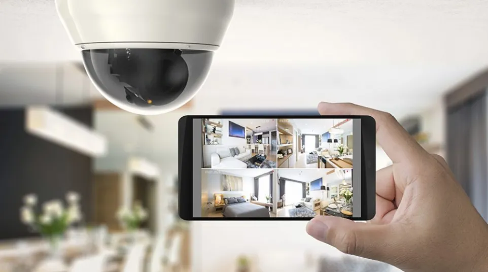 Smartphone displaying home security camera footage indoors under a dome camera capturing multiple room views