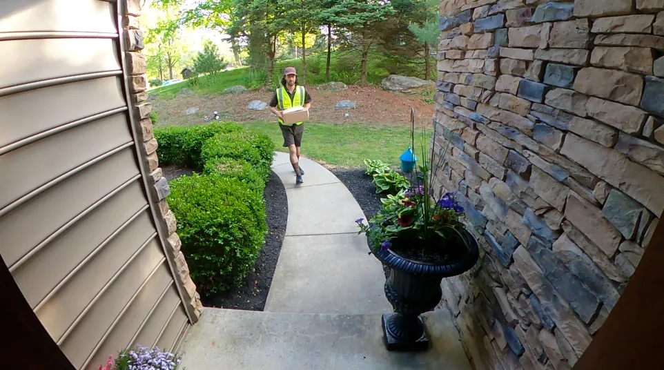 Delivery person in safety vest walking on a curved path carrying a package toward a house entrance