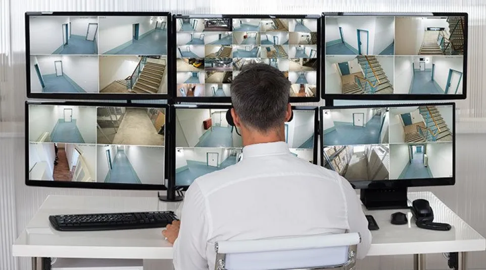 Man monitoring multiple security camera feeds on several screens showing building interiors and stairwells.