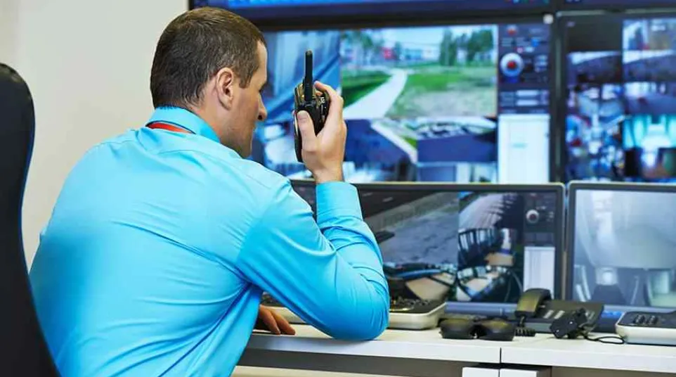 Security guard monitors multiple surveillance screens while communicating through a walkie-talkie in control room