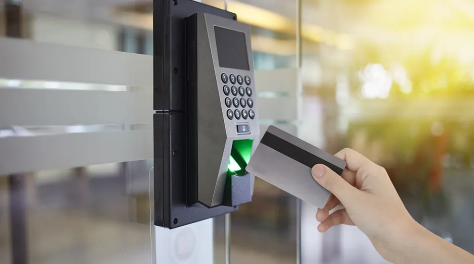 Hand holding access card near fingerprint scanner and keypad on glass door system with sunlight in background