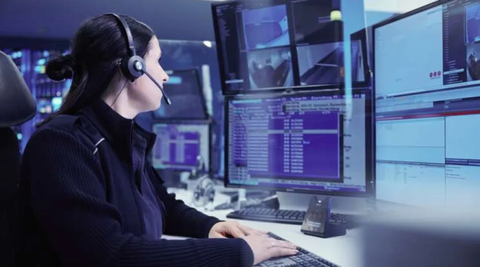Woman with headset monitoring multiple computer screens with code and surveillance footage in control room.