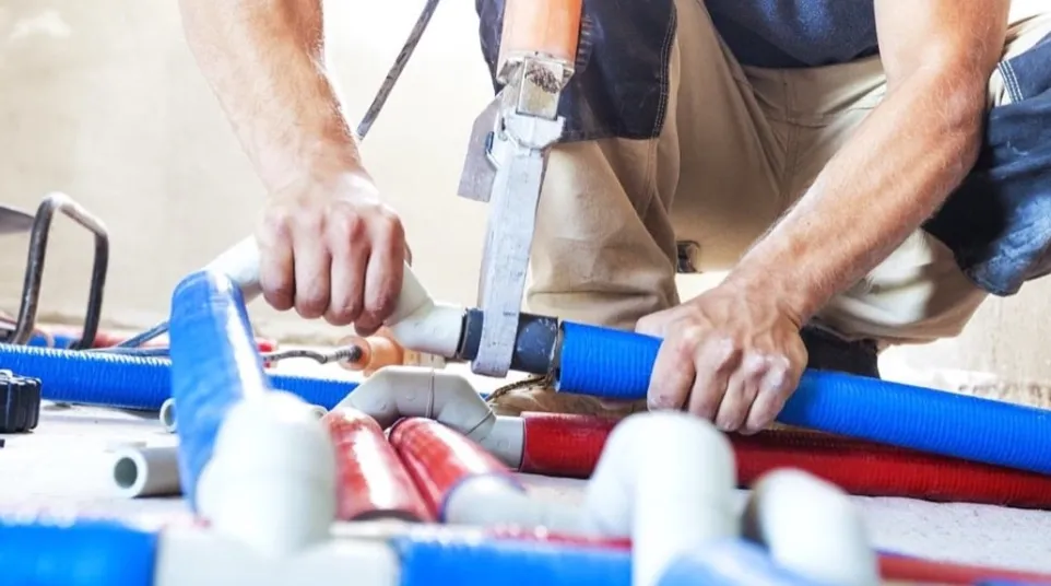 Plumber working with blue and red pipes using a cutting tool on a concrete floor in construction site.