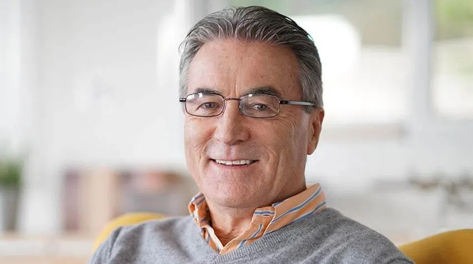 Smiling middle-aged man wearing glasses and a gray sweater sitting indoors in a bright room.