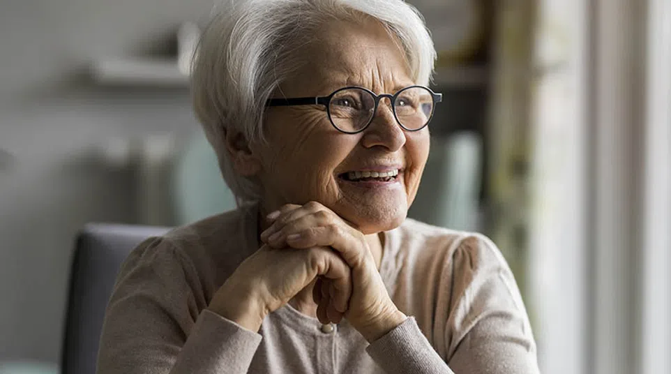Smiling elderly woman with white hair and glasses sitting indoors, hands clasped, looking out a window.