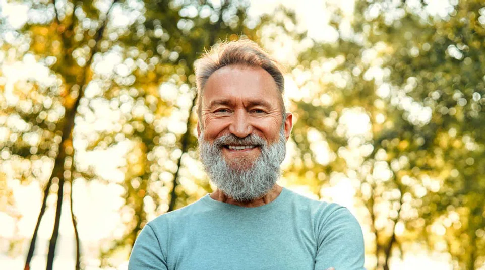 Smiling middle-aged man with gray beard and folded arms standing outdoors in a sunlit green park.