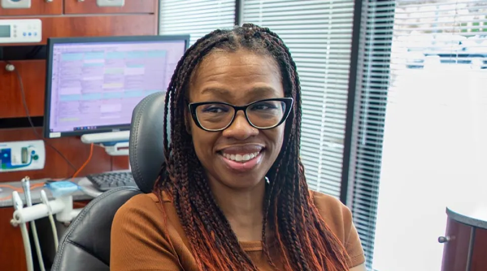 Smiling woman with glasses and braided hair sitting in a dental office chair near computer and equipment.
