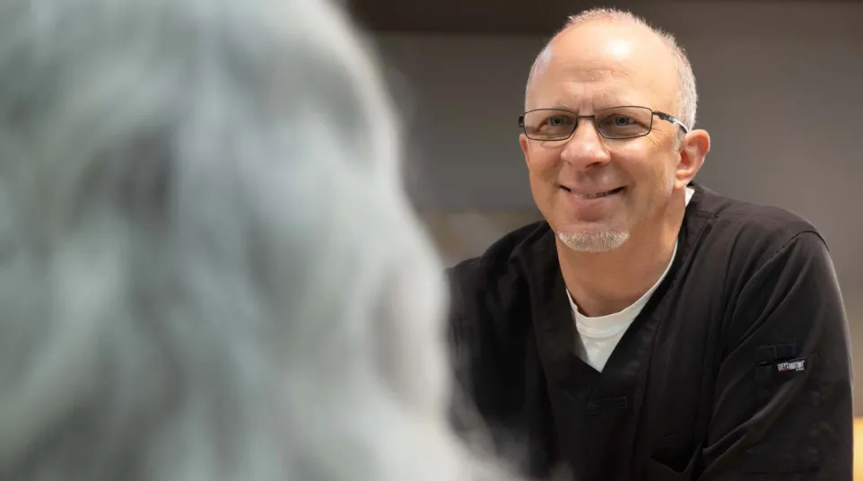 Smiling male healthcare professional wearing glasses and black scrubs talking to a patient with gray hair.