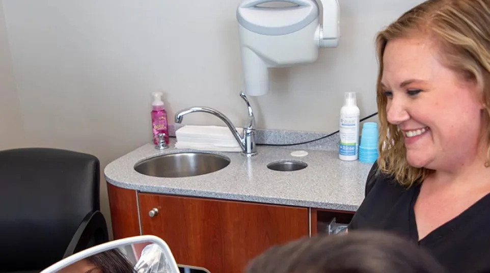 Smiling woman looks at her reflection in a hand mirror while a healthcare professional watches with a smile.