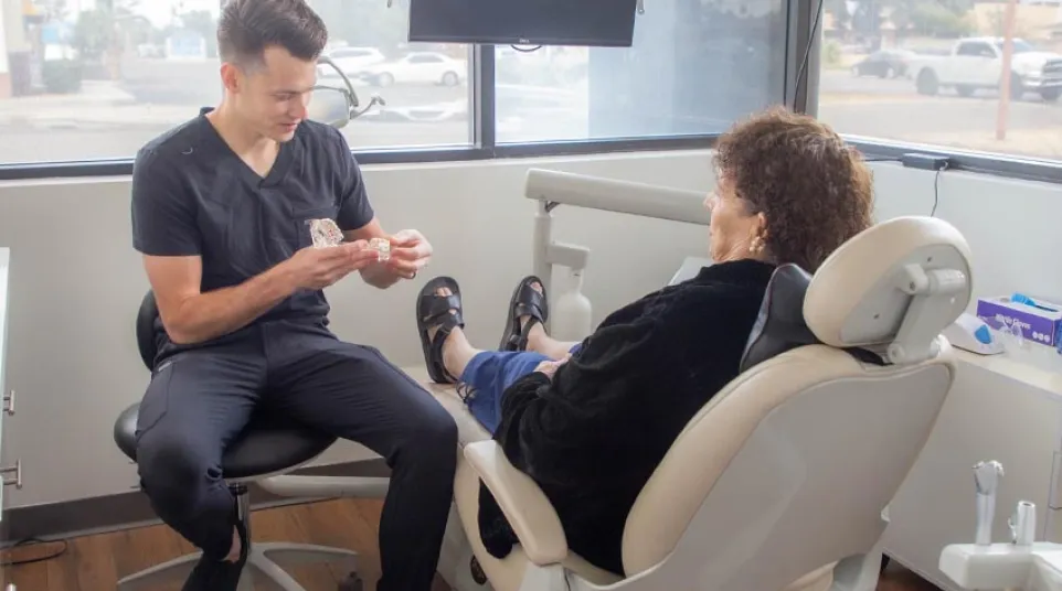 Dentist explaining dental mold to senior woman in modern dental office with large windows.
