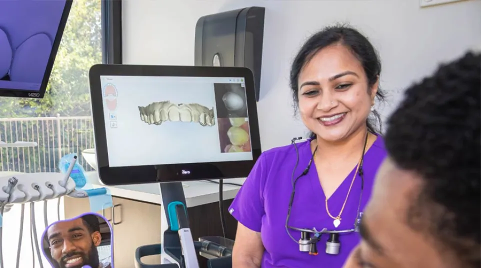Dentist in purple scrubs smiling at patient holding mirror showing his teeth and dental 3D scan on screen