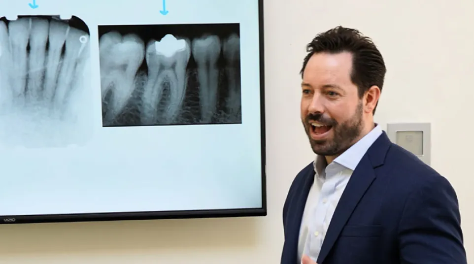 Dentist in suit explaining dental X-rays of teeth displayed on screen in a clinic setting.