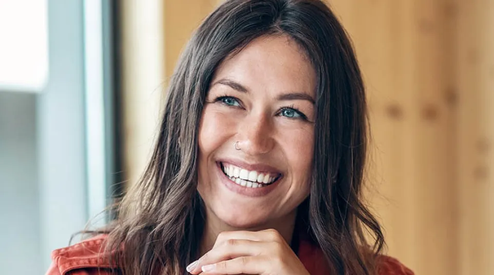 Smiling young woman with blue eyes and brown hair wearing red jacket sitting indoors by window.