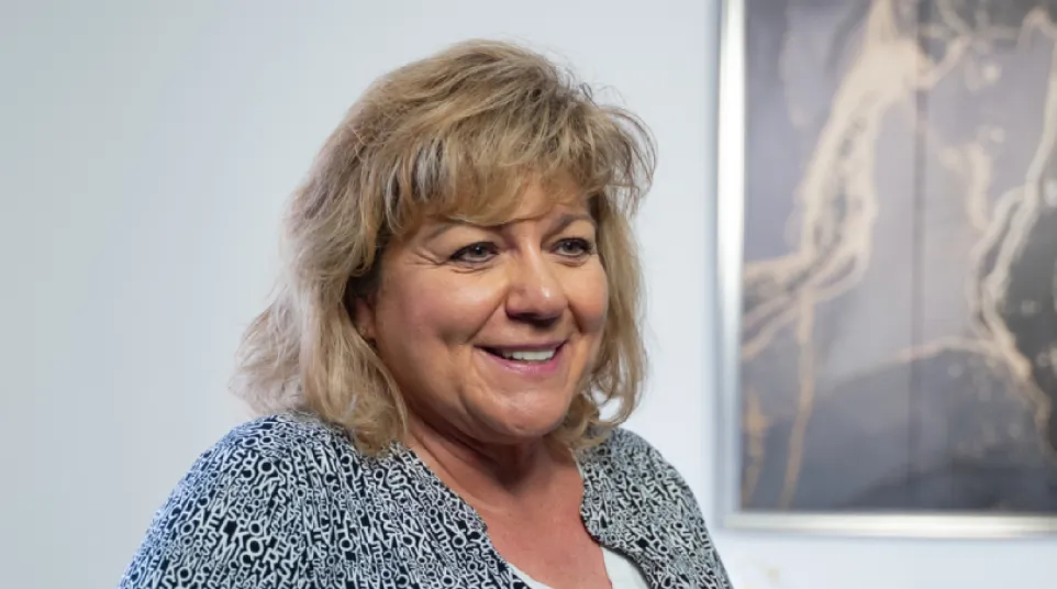 Smiling middle-aged woman in patterned jacket standing indoors with framed art in the background.