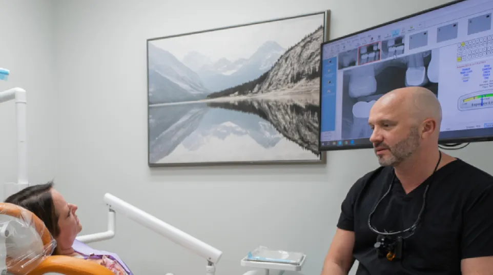Dentist consulting female patient in dental office with X-ray images on screen and mountain art on wall