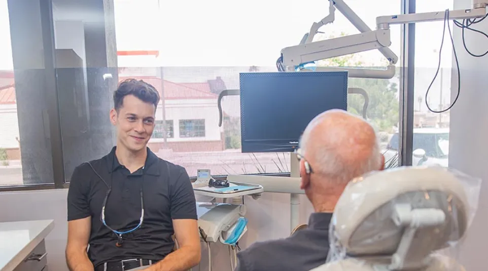 Young male dentist consults seated elderly patient in modern dental office with equipment and large window.