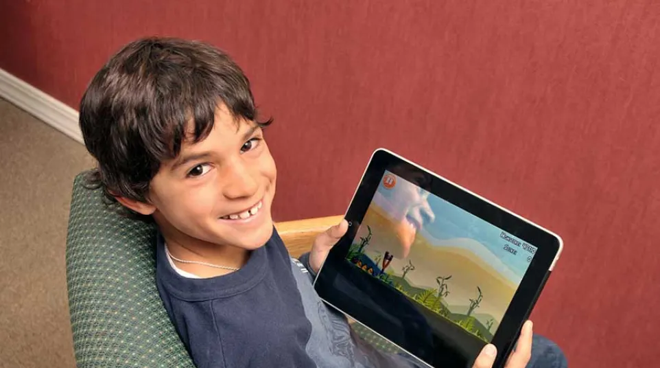 Smiling boy sitting in a chair holding a tablet displaying a colorful game with animated characters.