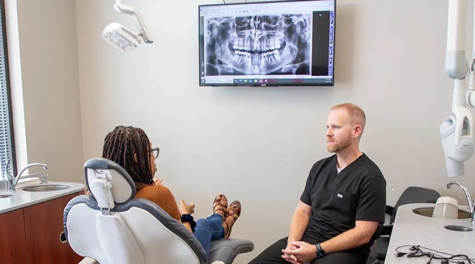 Dentist consulting patient in dental office with a large digital dental X-ray image displayed on the wall.