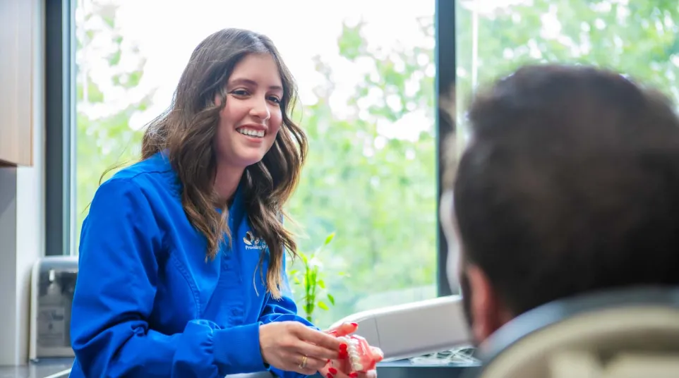 Smiling female dentist explaining dental model to patient in dental office with large window and greenery outside